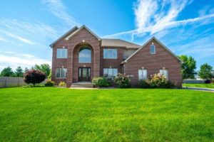 A large two-story brick house with an arched front door, represented by Crown Point Realtors, surrounded by a well-manicured lawn and a clear blue sky.