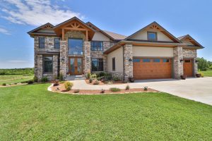 A modern two-story house with stone accents, wooden garage doors, and a landscaped front lawn under a clear blue sky, listed by Realtor Crown Point.