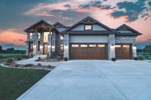Modern house with stone accents and large windows at dusk, driveway leading to a double garage under a colorful sky, listed by Crown Point Realtors.