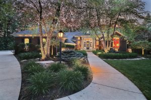 Illuminated pathway leading to a modern house with lighted windows at dusk, surrounded by lush trees and landscaped gardens in Crown Point.