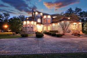 Large brick house with illuminated windows at dusk, featuring a cobblestone driveway and manicured bushes in the foreground, listed by Crown Point Realtors.