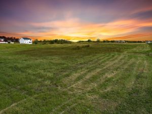 A scenic sunset over a rural landscape with lush green fields and trails, featuring a white house on the left and distant buildings, perfect for Realtors Northwest Indiana.