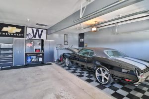 A neatly organized garage with a black vintage car, checkered floor, and tool cabinets adorned with automotive brand logos, listed by Crown Point Realtors.