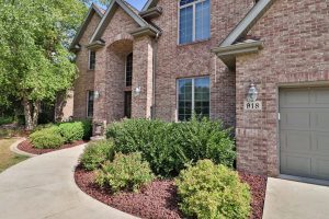 Two-story brick house with arched windows, a single garage door, and a landscaped front yard featuring shrubs and a concrete walkway offered by Crown Point Realtors.