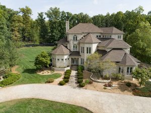Aerial view of a large, luxurious multi-story house with a shingled roof, represented by Crown Point Realtors, surrounded by landscaped gardens and trees, featuring a circular driveway.