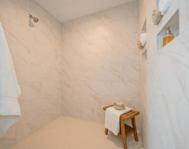 Spacious walk-in shower with light marble tile, a wall-mounted showerhead, built-in shelves, a wooden stool, and a white towel—a perfect touch found in many Valpo homes for sale.